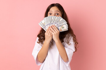 Portrait of surprised little girl wearing white T-shirt covering half of face with big fan of dollar banknotes, looking at camera with big eyes. Indoor studio shot isolated on pink background.