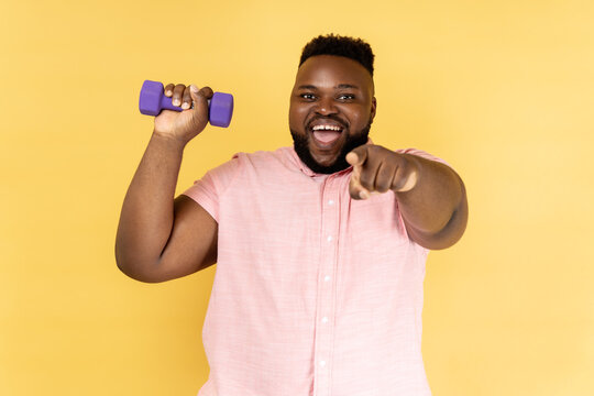 Portrait of cheerful joyful man wearing pink shirt standing with dumbbell and pointing to camera, inviting you to fitness club. Indoor studio shot isolated on yellow background.