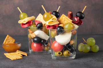 Jarcuterie in two glass includes various types of sausages, pickles, cheese, nuts, vegetables and fruits. Modern version of festive snack on brown background
