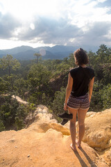 Caucasian woman, sitting on the edge of a cliff appreciating the landscape. Mountains and trees are seen.