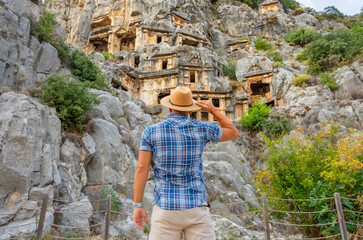 Swarthy caucasian european guy in plaid shirt and a hat stands back and looks in surprise at the ancient unique famous places Turkey, Antalya Demre, Myra, ancient lycian tomb casts faces.