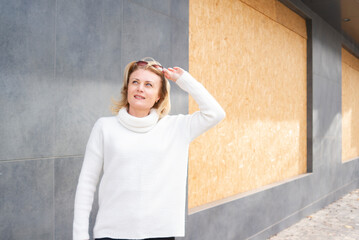 Blonde woman in white sweater in front of a boarded up window for security. Middle-aged woman on a walk in the city. Woman urban lifestyle. Dreamy European lady concentrated somewhere. 