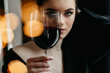 Portrait of serious woman looking at camera through glass of red wine. Stylish woman in black outfit, holding glass of drink and posing on background of bokeh light