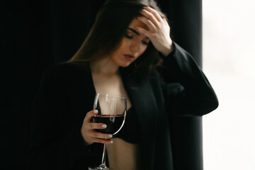 Blurred of confident woman in formal suit holding glass of drink. Adult girl, wearing in black jacket and bra, looking down and holding glass of red wine, standing in front of curtains in dark room