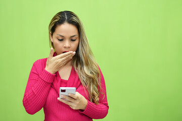 Mujer Joven Latina sorprendida mientras ve su telefono movil. Aislada sobre fondo verde, espacio...