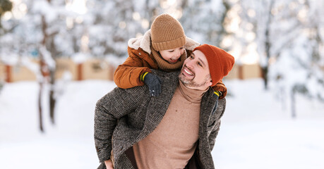Dad giving piggyback ride to happy little son while walking outdoors in snowy winter park
