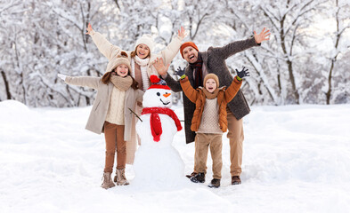 Happy family in warm clothes laughing merrily and raising hands up while making snowman together
