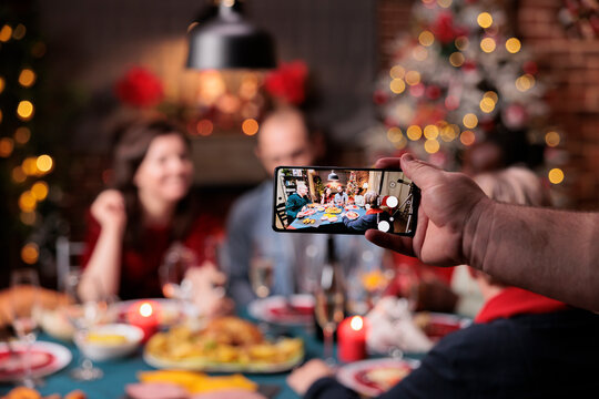 Diverse family celebrating christmas, focus on smartphone photo, friends eating at festive dinner table on blurred background. Xmas holidays celebration, hand holding mobile phone