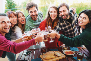 Group of cheerful young friends celebrating with wine and beer glasses at picnic happy hour party in the terrace - Young people having fun drinking and eating outdoor - Friendship and youth lifestyle