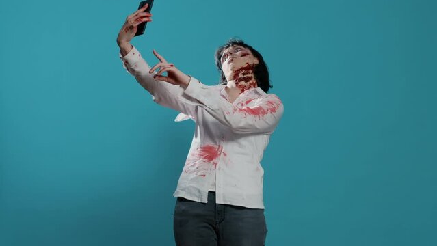 Undead walking corpse taking selfie photo with smartphone device on blue background. Spooky and scary looking woman moster with deep and bloody wounds and scars taking photo of herself.