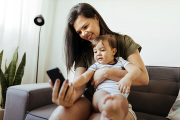Mother and baby playing with a smart phone sitting on a couch in the living room at home