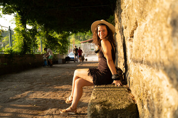 Beautiful young caucasic woman with a hat sitting in a stone bench in the streets of spain - obrazy, fototapety, plakaty