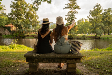 two friend girls with hats chatting sitting on a stone bench in front of the river