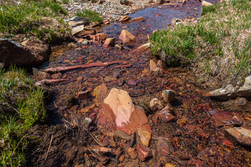 Sharp stone in clear water stream with motley stony bottom in sunlight. Colorful mountain stream with mineral water in bright sun. Picturesque top view to sunlit multicolor stones in spring water.