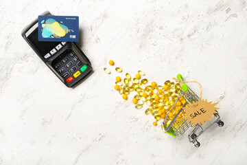 Shopping cart with fish oil capsules, payment terminal, credit card and tag with word SALE on light background