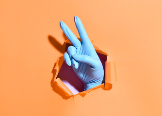 Hand in medical glove showing victory gesture through hole in paper on color background