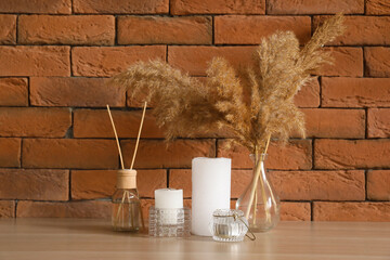 Candles, reed diffuser and vase with pampas grass on table near brick wall
