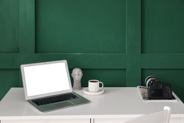 Professional photo camera, magazine, cup of coffee and laptop on table near green wall
