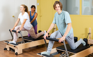 Teenagers boy and girl practicing Pilates system on reformer supervised in gym
