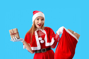 Happy young woman in Santa costume with bag of gifts on blue background