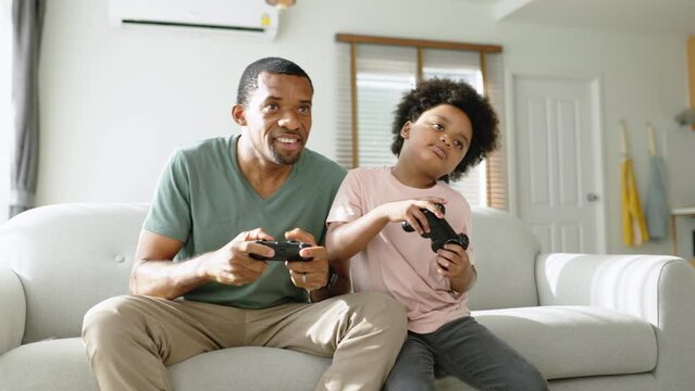 Black African American Father and his Little son sitting on couch using game controllers playing console video game