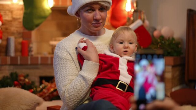 Beautiful smiling woman posing with baby girl as friend taking photo on smartphone in slow motion. Portrait of happy Caucasian young aunt and toddler niece celebrating New Year's eve at home