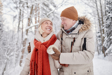 Waist up portrait of happy senior couple enjoying walk in winter forest and looking at each other with love