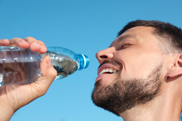 Happy man drinking water against blue sky on hot summer day, closeup. Refreshing drink