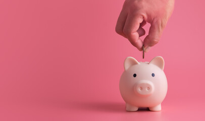 Hand putting coins in pink pig piggy bank on pink background with copy space, investment saving money wealth and financial accounts concept.