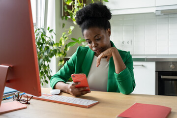 Focused African American woman holding phone reading business press on mobile apps sits at computer desk in home office. Brazilian female uses smartphone to send e-mail mail to company employees