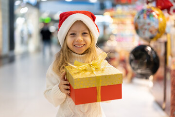 Fototapeta na wymiar a little girl in a Christmas hat with a New Year's gift in her hands at a showcase with toys. children choose gifts for Christmas. Black Friday discounts