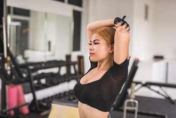 A slim and sexy asian woman in a black crop top does overhead tricep stretches before a workout session at the gym.