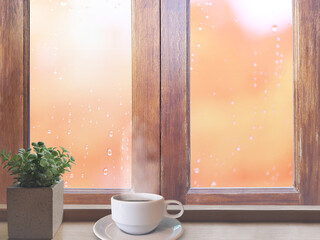 On a rainy day, water droplets were visible on the outside glass blurred. (Window background)
On the table, place a white cup of coffee and Small green leaves in pot.