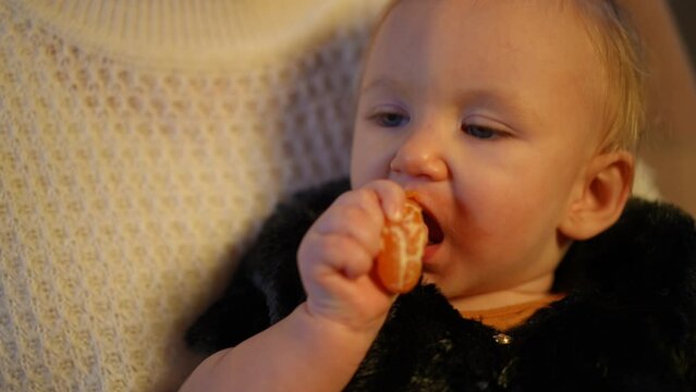 Headshot portrait of toothless charming baby girl sucking mandarin sitting in hands of unrecognizable woman indoors. Close-up of cute Caucasian child enjoying tasty healthful fruit