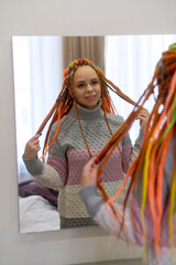 Woman fixing colourful hairstyle in mirror. Positive young female hipster in casual clothes looking in mirror and touching long colorful dreadlocks gently at home