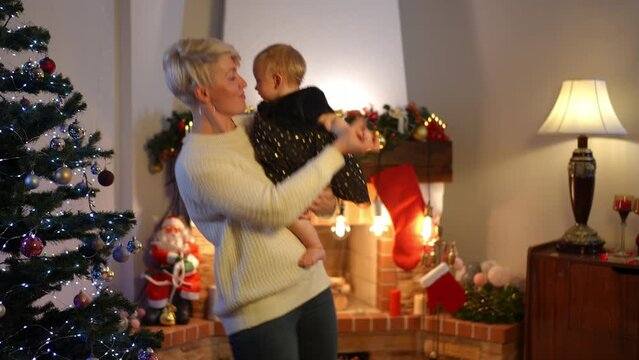 Side view happy woman dancing with baby girl in living room on Christmas eve. Portrait of positive relaxed Caucasian aunt and niece having fun at home indoors on New Year smiling