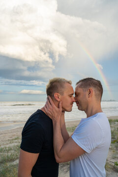 gay couple embracing with hand around neck with rainbow at beach 