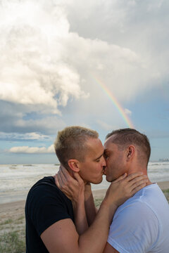 gay couple kissing with hands around neck with rainbow at beach 