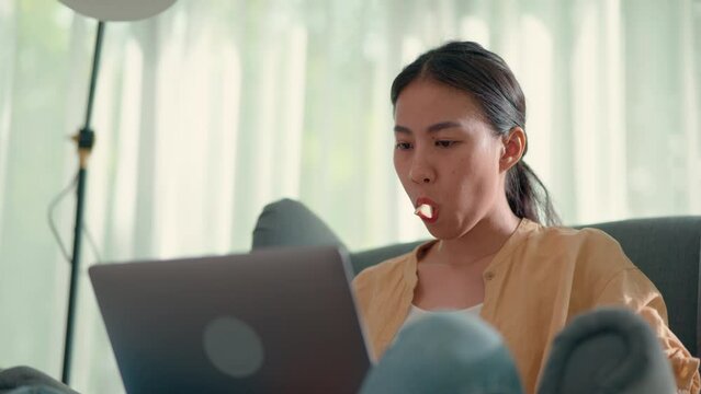 Young pretty woman sitting on sofa and eating apple while typing in laptop computer, new normal and working from home concept