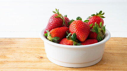 Close view of a white ceramic bowl with red fresh strawberries inside  placed on top of a wooden table 