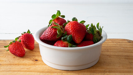 close view of a white ceramic bowl with red fresh strawberries inside and two of them placed on top of a wooden table 