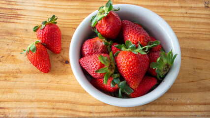 Flat lay view of a white ceramic bowl with red fresh strawberries inside and two of them placed on top of a wooden table