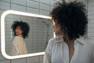 Young woman with dark curly hair checking clothes in the mirror in modern washroom.