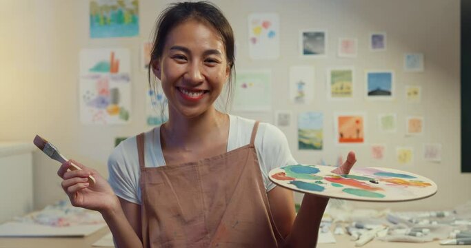 Close-up Happy cheerful youth Asian Female wear apron smile look at camera hold palette of color and brush with painting artwork cozy workshop in home at night. Painter art creativity concept.