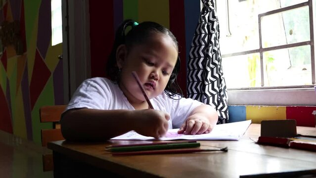 Cute asian little girl sitting near window while coloring her book. Kindergarten kid doing her homework at home