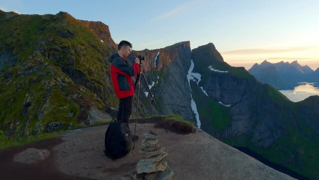 Medium shot of a man standing on the famous peak of reinbringen in Lofoten Norway, putting a camera on a tripod with magnificent views of rocky peaks in the background