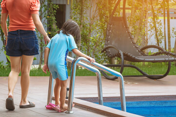 Adorable daughter in blue swimming suit going down swimming pool ladder to swim with clear blue water and her mother to take care of closely. Little girl at pool. Happy family relationship at pool.