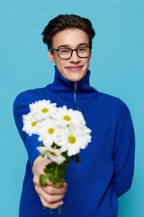 vertical portrait of a cute man in black eyeglasses and a blue zip-up sweater, holding out a bouquet of white daisies to the camera with a wide smile on his face