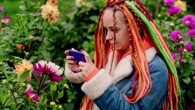 Woman taking pictures on smartphone in park. Positive young female hipster with long dreadlocks in casual clothes photographing flowers on mobile phone in park