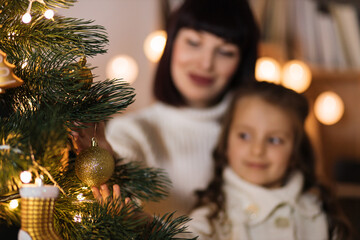 Focus on Christmas toy in shape of gold ball on decorated tree. Happy small preschool caucasian child girl decorating tree with young mother, enjoying preparing for New Year celebration at home